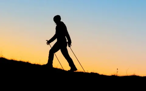 Sylwetka osoby uprawiającej nordic walking na tle zachodzącego słońca. Czy wolisz nordic walking rano czy wieczorem?