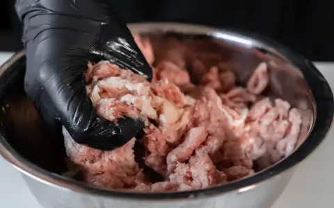 Ręka w czarnej rękawiczce miesza mięso mielone w metalowej misce, przygotowując obłędne kotlety mielone.
