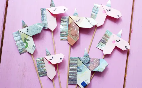 Kreatives Geld falten: Mehrere Einhorn-Figuren aus Geldscheinen und Glitzerpapier auf Holzstäbchen vor rosafarbenem Hintergrund.