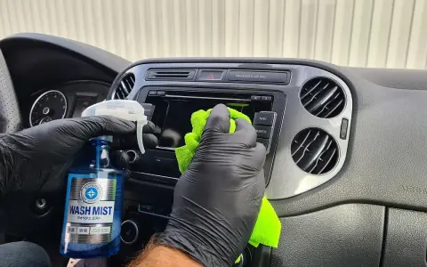 Ręce w czarnych rękawiczkach czyszczą deskę rozdzielczą samochodu. Pokazano, jak wypolerować plastik domowym sposobem, używając płynu "Wash Mist".