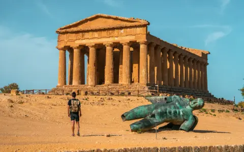 A lone traveler explores the Valley of the Temples in Sicily, standing before an ancient Greek temple and a fallen bronze statue.
