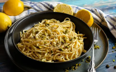 Eine Schüssel mit Zitronen Parmesan Pasta, garniert mit gehackten Pistazien und schwarzem Pfeffer. Frische Zitronen liegen daneben.