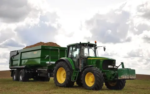 Zielony traktor John Deere z przyczepą wypełnioną ziemniakami na polu.