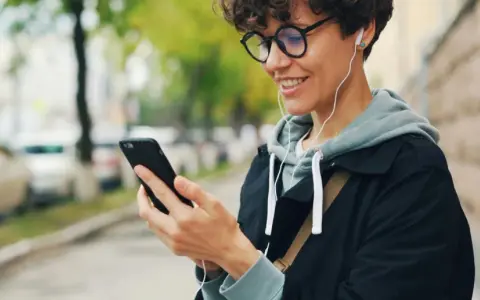 Uśmiechnięta osoba w słuchawkach patrzy na telefon. Czy wiesz, co to jest impedancja w słuchawkach i jak wpływa na dźwięk?