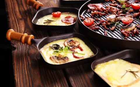 Raclette co to? Roztopiony ser z dodatkami w małych patelenkach i grillowane mięso z pomidorkami na drewnianym stole.