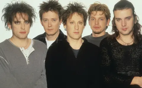 The Cure członkowie w szarych i czarnych ubraniach, z charakterystycznymi fryzurami i makijażem.