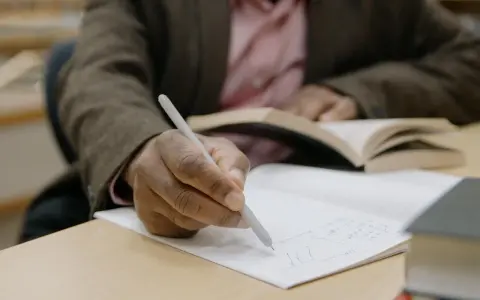 Osoba pisze ręcznie notatki w zeszycie, przygotowując list motywacyjny erasmus. W tle widać otwartą książkę.