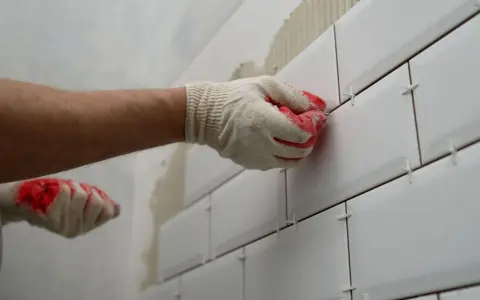 Rękawica w białej rękawicy z czerwonymi akcentami układa białe płytki na ścianie, pokazując, jak przygotować ścianę pod płytki.