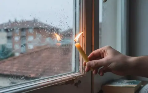 Una vela encendida cerca de una ventana con gotas de lluvia. El humo de la vela se arremolina, y parece que entra aire por la ventana, reflejando edificios en el exterior.
