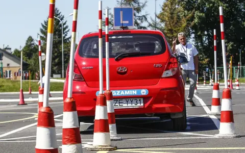 Czerwony Hyundai na placu manewrowym, z literą "L" na dachu. Instruktor pokazuje znak pokoju. Przygotowania do egzaminu na prawo jazdy b+e.