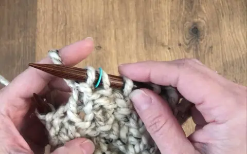 Manos tejiendo con agujas de madera un punto de relieve con lana gruesa y un marcador azul.