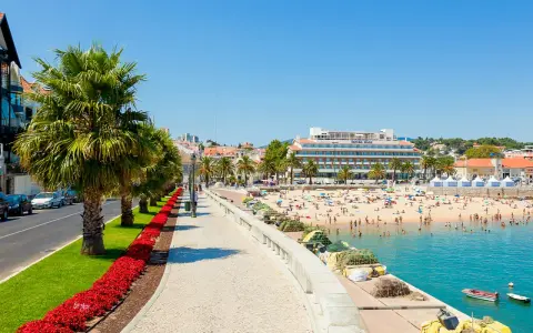 Sonniger Strandtag in Cascais bei Lissabon. Palmen säumen die Promenade, Menschen genießen das Meer und das Hotel Baía ragt empor.