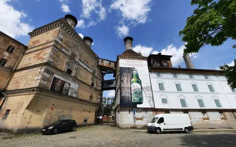 Stara fabryka z wielkimi plakatami reklamującymi piwo Namysłów. Biała furgonetka i czarny samochód stoją na brukowanej ulicy.