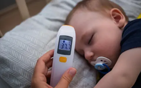 Elektroniczny termometr pokazuje 37.7°C, mierząc temperaturę niemowlaka śpiącego ze smoczkiem.