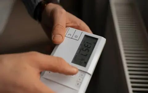 Dłoń naciska przycisk na białym sterowniku, by ustawić temperaturę na grzejniku.