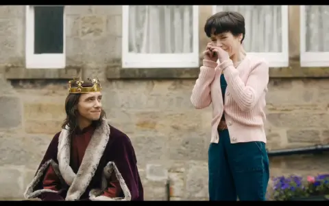 Un re con la corona e la pelliccia, forse il protagonista di "the lost king film", guarda una donna che ride con le mani sul viso.