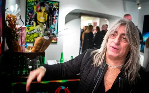 Mikkey Dee son, en man med långt grått hår och en silverfärgad kedja, poserar vid ett bord med blommor och flaskor.