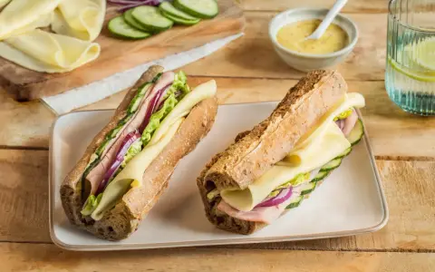 Deux sandwichs jambon fromage appétissants, garnis de concombre, oignon rouge et laitue, sur un plateau blanc.