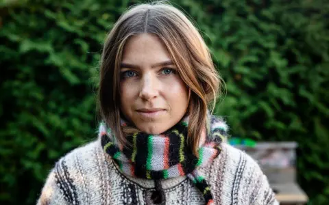 Elin Berlin med blå ögon och fräknar, klädd i en stickad tröja och randig halsduk, står framför en grön häck.