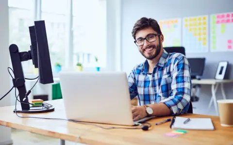 Uśmiechnięty mężczyzna w okularach pracuje przy laptopie. Cieszy się, bo właśnie dostał umowę na czas nieokreślony.