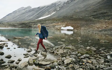 Kobieta z plecakiem przemierza kamieniste brzegi górskiego jeziora. W tle skaliste zbocza, idealne dla skandynawskie marki outdoorowe.
