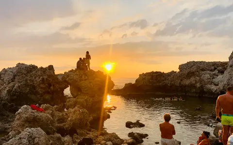 Tramonto dorato sulla piscina di Venere a Milazzo. Persone sedute sulle rocce e in acqua, godendosi la vista.