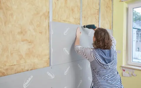 Frau verkleidet Wand mit Rigipsplatten und Schrauber. Isolierung ist sichtbar.