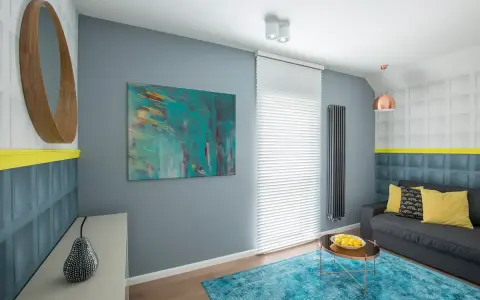 Sala de estar con sofá gris, alfombra turquesa y arte abstracto en tonos fríos. Un espejo redondo de madera y detalles en amarillo complementan el espacio.