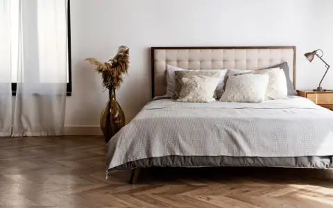 Cama con base tapizada o canapé, cojines mullidos y mantas grises. Un jarrón con pampas decora el rincón.