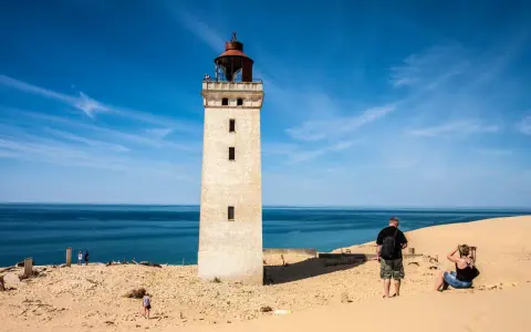 Ett vitt utsiktstorn i Danmark reser sig mot en blå himmel. Sanddyner och hav omger tornet.