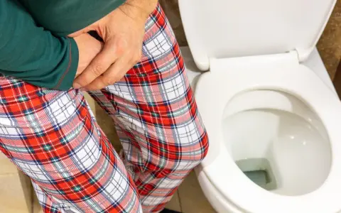 Mężczyzna w piżamie z kratę trzyma się za krocze, siedząc obok toalety. Czy zwapnienie prostaty to rak?