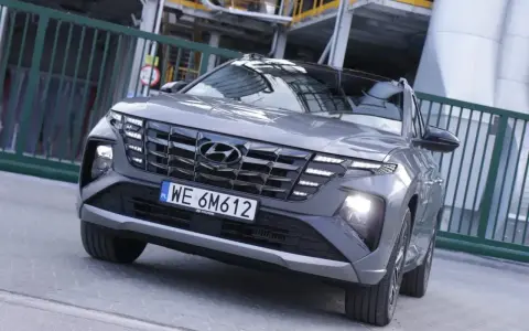 Szary Hyundai Tucson hybryda z charakterystycznym grillem i światłami LED, zaparkowany przed zielonym ogrodzeniem.