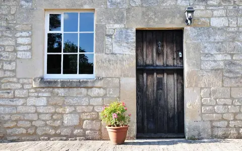 Facciate in pietra e mattoni grezzi con una porta in legno scuro e una finestra bianca. Un vaso di fiori rosa aggiunge un tocco di colore.