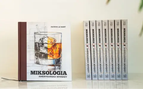 Książka "Miksologia Amerykańska Whiskey" Patryka Le Narta, z ilustracją szklanki z lodem i bursztynowym trunkiem. Obok niej rząd identycznych tomów.