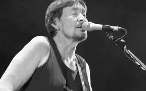 Chris Rea nie żyje. Muzyk śpiewa do mikrofonu na czarnym tle, w czarnej koszulce bez rękawów.