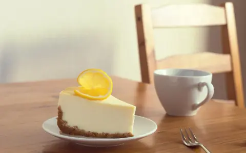 Une part de le meilleur cheesecake au citron, garnie d'une tranche de citron, sur une assiette blanche, à côté d'une tasse et d'une fourchette sur une table en bois.