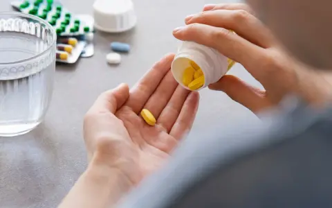Dłoń wysypująca żółte tabletki z pojemnika na drugą dłoń, obok szklanka wody i opakowania leków.