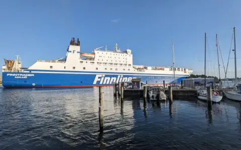 Duży prom Finnlines "Finntrader Malmö" cumuje w porcie, gotowy do rejsu. W tle widać żaglówki. Prom do Finlandii czeka na pasażerów.