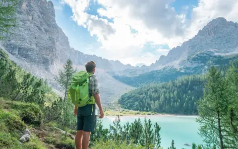 Wanderer mit Rucksack blickt auf türkisfarbenen Bergsee. Die Alpenlandschaft erinnert an die Grenze zwischen Schweiz und Italien.