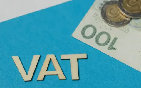 Drewniane litery VAT na niebieskim tle, obok banknot 100 zł i monety. Symbolizuje zwolnienie z VAT.