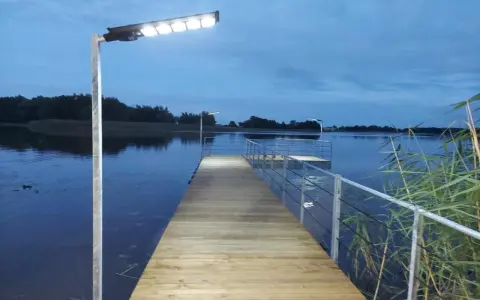 Drewniany pomost nad jeziorem, oświetlony lampami, zachęca do spaceru. Czy pomost nad jeziorem może być prywatny? Ten wygląda na dostępny dla wszystkich.