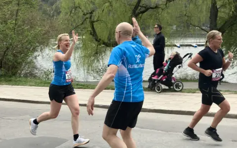 Två löpare ger varandra high-five under roliga lopp. En kvinna i blått och en man i blått. I bakgrunden syns en barnvagn och en sjö.
