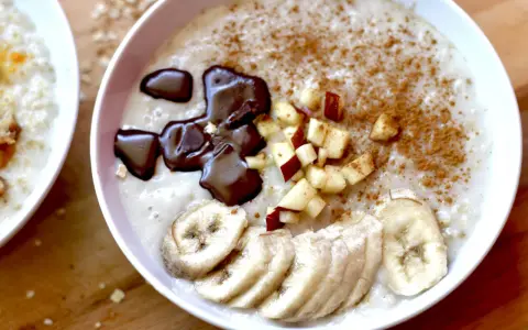 Un tazón de avena cremosa, perfecto para aprender como hacer porridge. Decorado con rodajas de plátano, trocitos de manzana, canela y un toque de chocolate derretido.