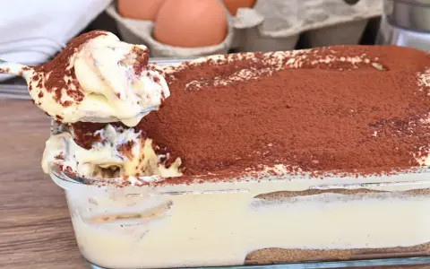 Un cucchiaio preleva una porzione cremosa di tiramisù, con 500 g di mascarpone, cosparso di cacao. Sullo sfondo, uova fresche.