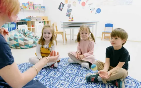 Dzieci siedzą na dywanie, słuchając nauczycielki. To przykład metody dobrego startu, gdzie nauka odbywa się przez zabawę.