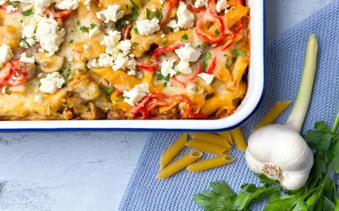 Herzhafter Gyros Auflauf mit Penne, Paprika und Feta, garniert mit Petersilie. Daneben liegen Penne, Knoblauch und frische Kräuter.