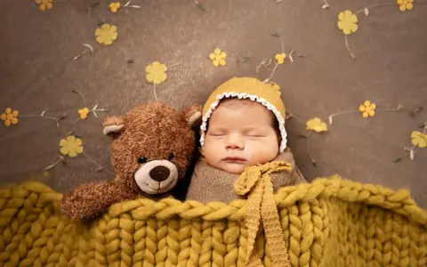 Bambino addormentato con orsacchiotto, avvolto in una coperta gialla. Un'idea per foto neonati fai da te con decorazioni floreali.