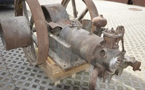 Zardzewiały silnik na gaz drzewny, z dużymi kołami, stoi na drewnianych klockach.