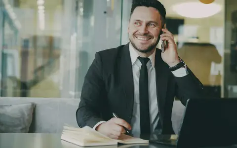 Uśmiechnięty mężczyzna w garniturze rozmawia przez telefon, notując coś w zeszycie. To właśnie taki mikroprzedsiębiorca buduje swój biznes.