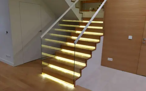 Nowoczesne schody dywanowe na beton z podświetleniem LED. Szklana balustrada i drewniane stopnie tworzą elegancką całość.